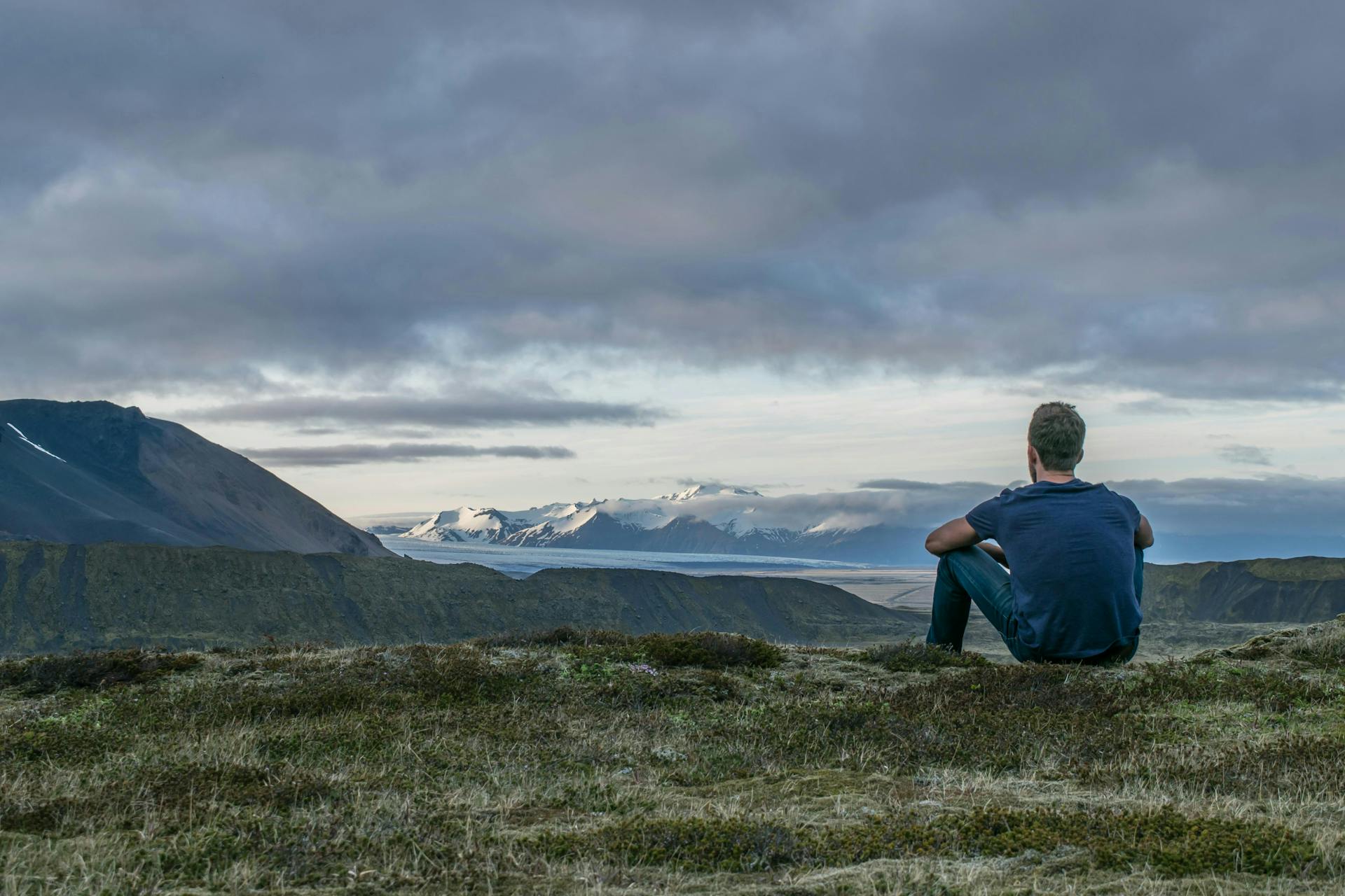 A man sitting on a hill that overlooks a fjord. Snowy mountains in the distance.